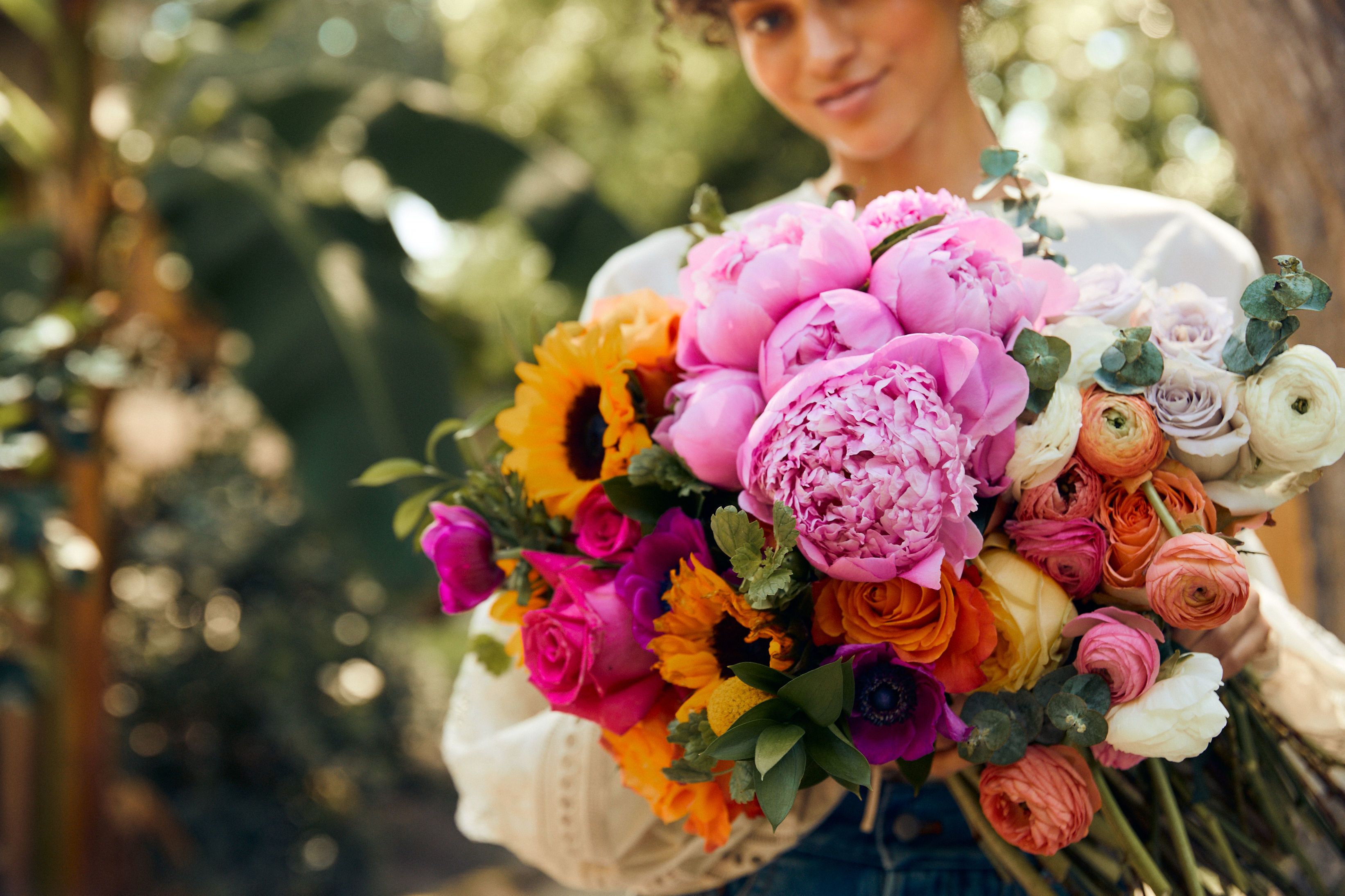 Close up of woman holding a sunflower, peonies, and roses bouquet.