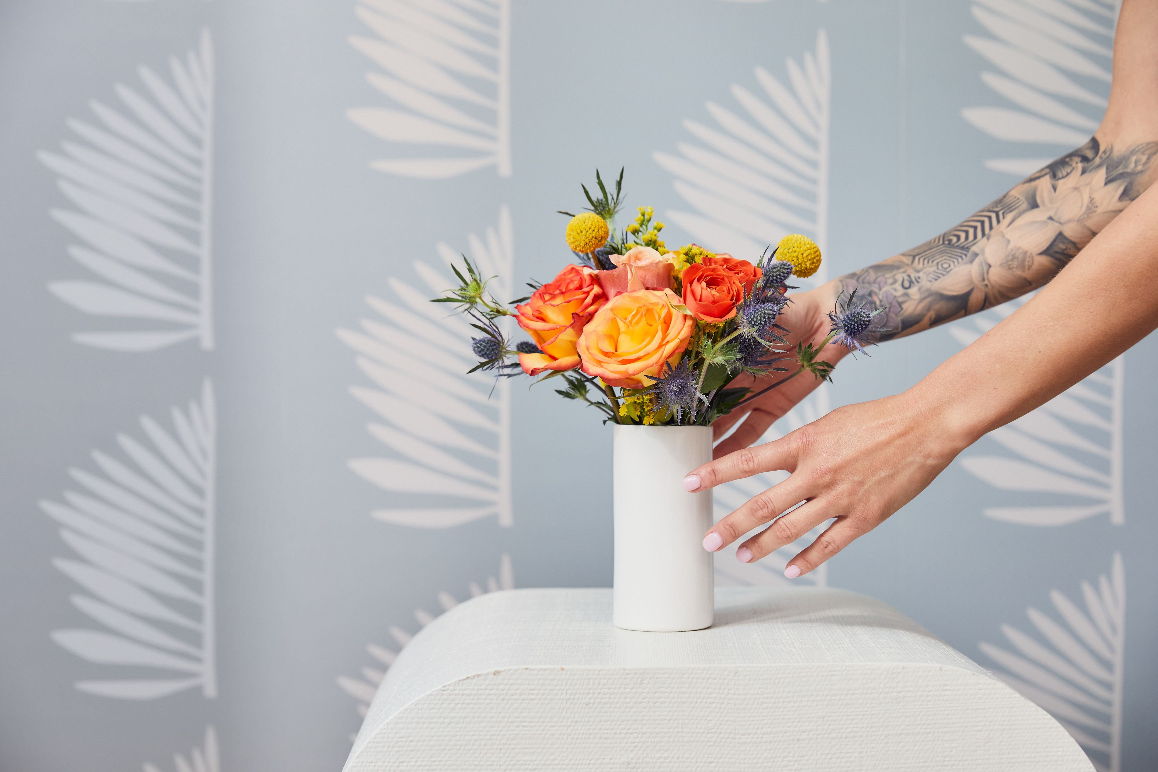 Close up of an arrangement in a white vase with a woman's hand.